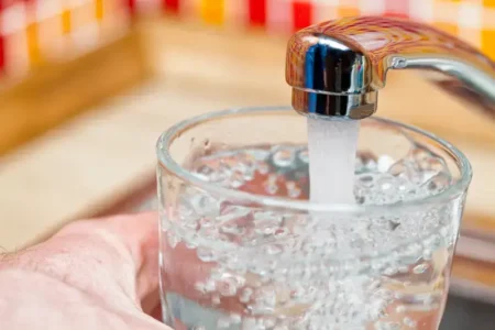 A person holds a clear glass under a kitchen faucet. Tap water pours from the faucet into the cup.